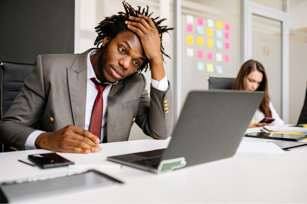 Mann sitzt am Schreibtisch vor einem Laptop, hält sich den Kopf und arbeitet im Büro, Kollegin im Hintergrund.